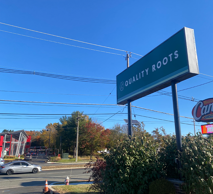 Quality Roots street sign in Marlton, New Jersey, standing tall against a clear blue sky with a busy road, nearby businesses, and autumn trees in the background